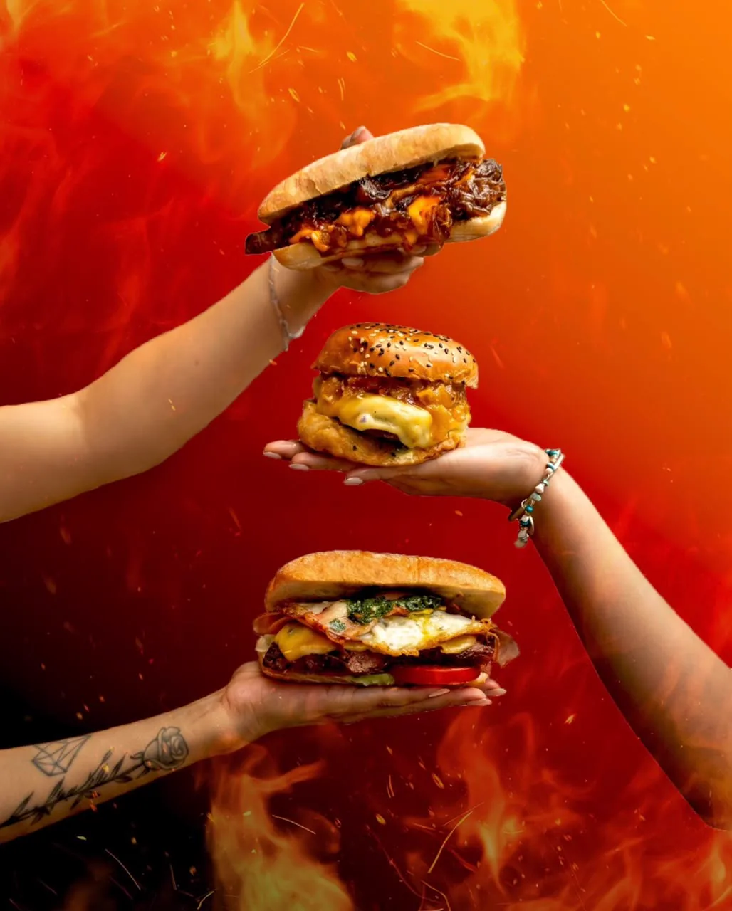Tres manos sosteniendo sánguches y hamburguesas de Pan y Fuego sobre un fondo de llamas